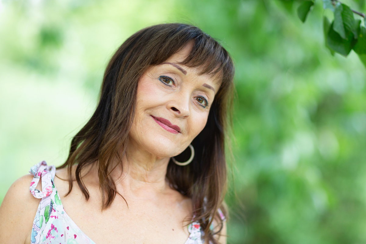 kobieta w wieku 50+ z ciemnymi długimi włosami z grzywką i z kolczykami oraz w makijażu stoi na zewnątrz w sukience i smutno się uśmiecha