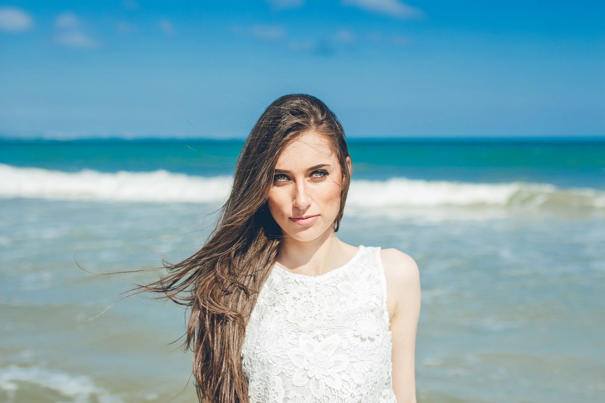 młoda dziewczyna w wieku około 30 lat z długimi włosami w białej koronkowej bluzce stoi na tle morza i lekko się uśmiecha