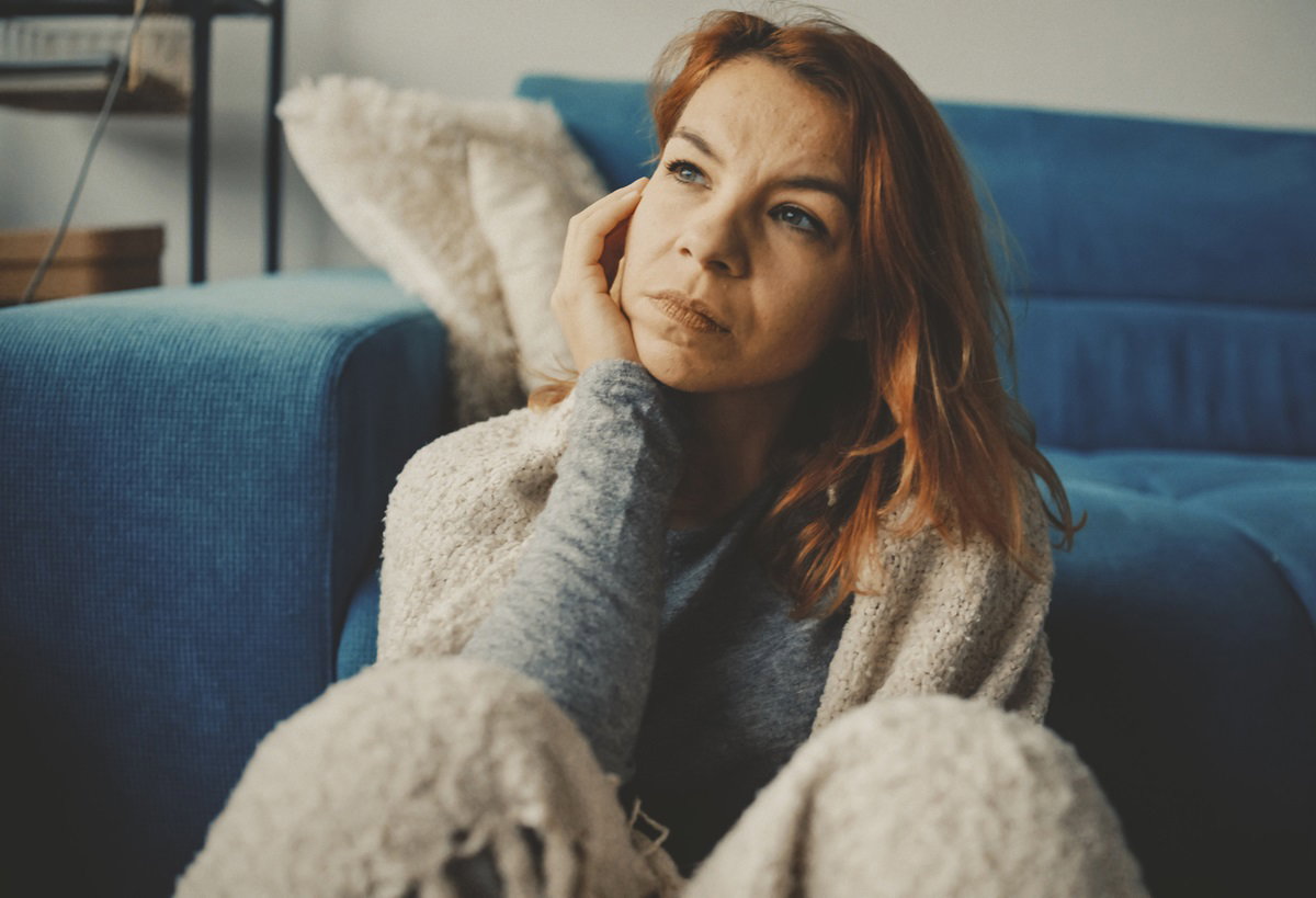 kobieta w wieku około 35 lat z rudymi włosami siedzi na podłodzie w salonie w kocu i rozmyśla