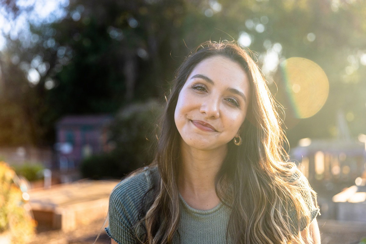 młoda kobieta w wieku około 30 lat z długimi ciemnymi włosami stoi na zewnątrz i uśmiecha się z przekąsem