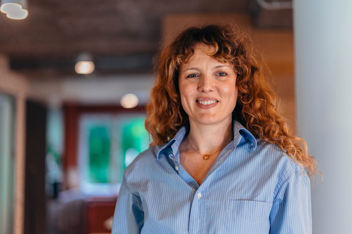 kobieta w wieku około 45 lat w niebieskiej koszuli w prążki z kręconymi rudymi włosami stoi u lekko się uśmiecha z przekąsem