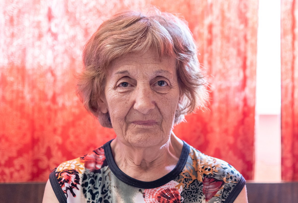 kobieta w senioralnym wieku 70+ z krótkimi włosami w kolorowej bluzce na tle czerwonej firanki siedzi zmartwiona