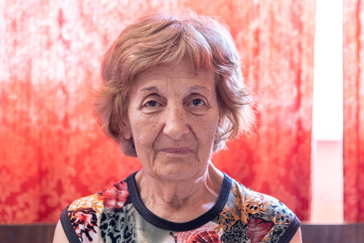 kobieta w senioralnym wieku 70+ z krótkimi włosami w kolorowej bluzce na tle czerwonej firanki siedzi zmartwiona