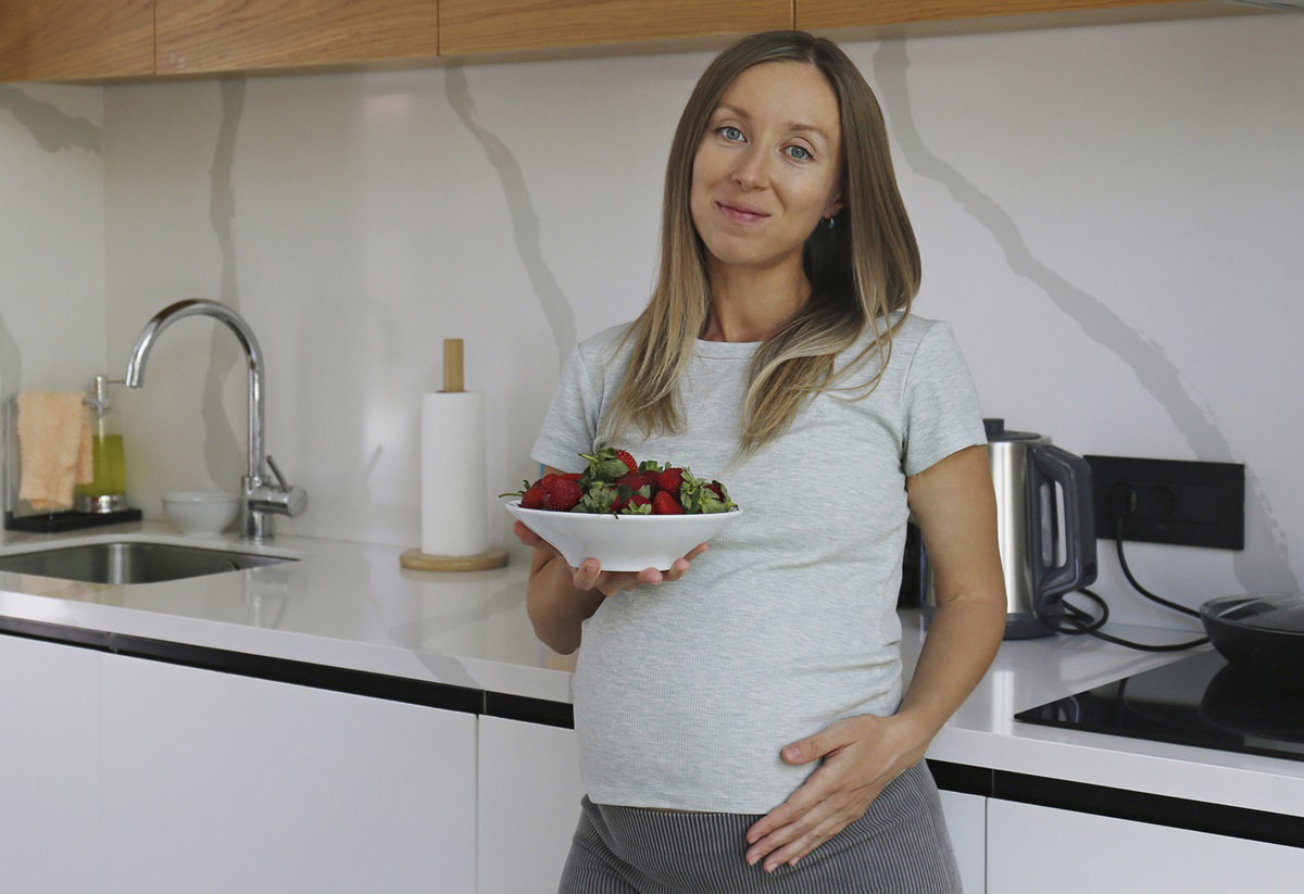 kobieta w wieku 30 plus w ciąży stoi w kuchni z miseczką truskawek i uśmiecha się nieśmiało