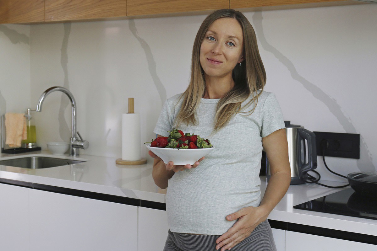 kobieta w wieku 30 plus w ciąży stoi w kuchni z miseczką truskawek i uśmiecha się nieśmiało