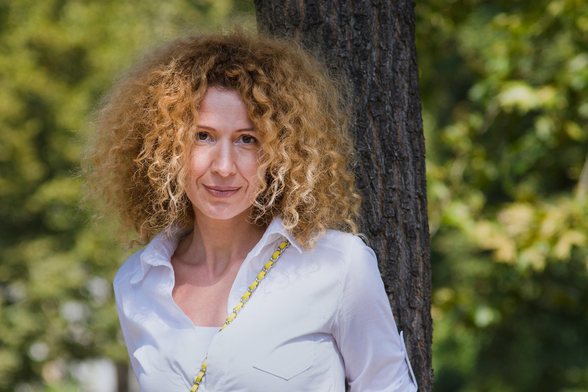 kobieta w średnim wieku około 40 plus z burzą loków na głowie opiera się o drzewo i aptrzy zamyślona