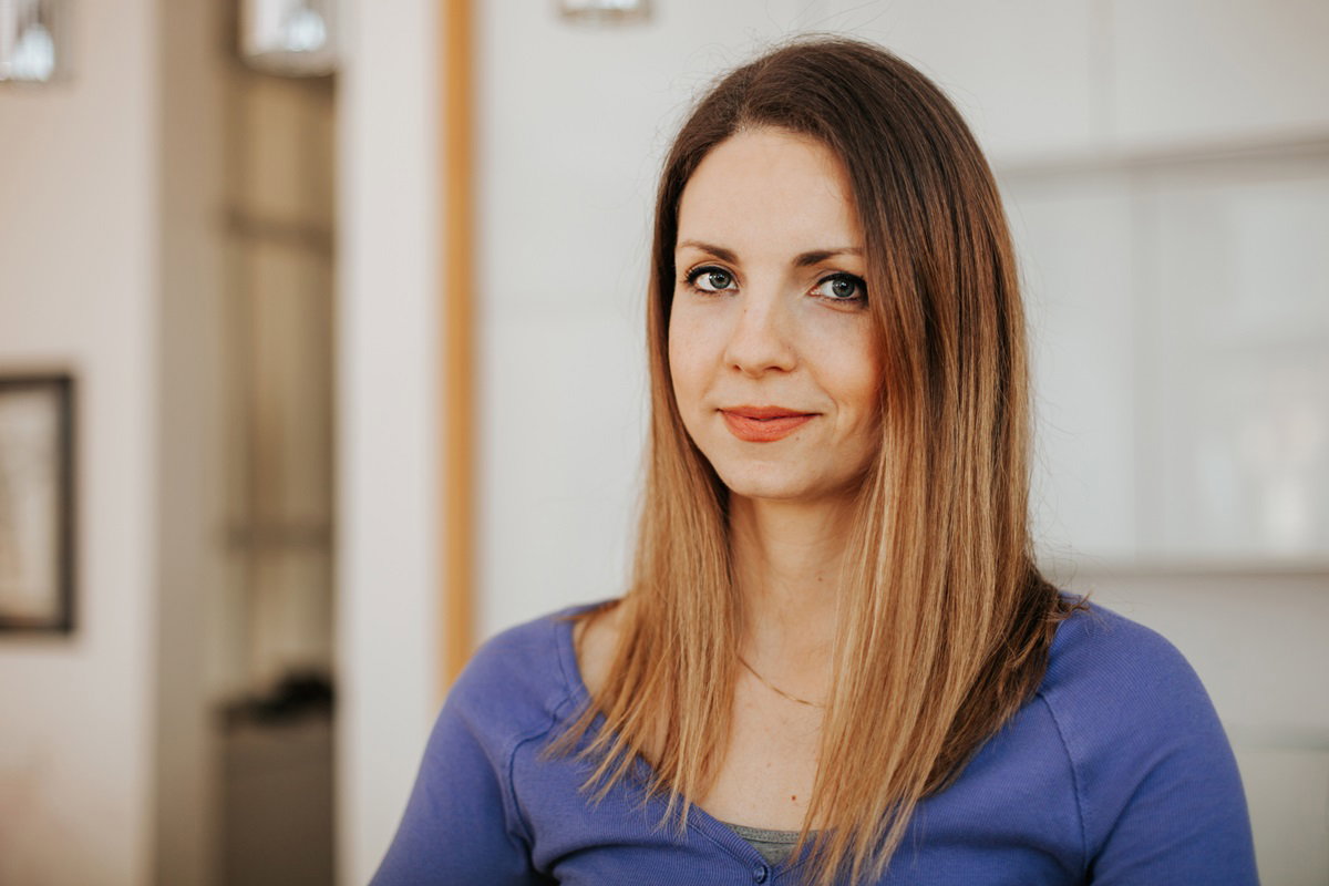 młoda kobieta w wieku około 30 lat z długimi włosami i w niebieskiej bluzce siedzi w salonie z niepewnym uśmiechem