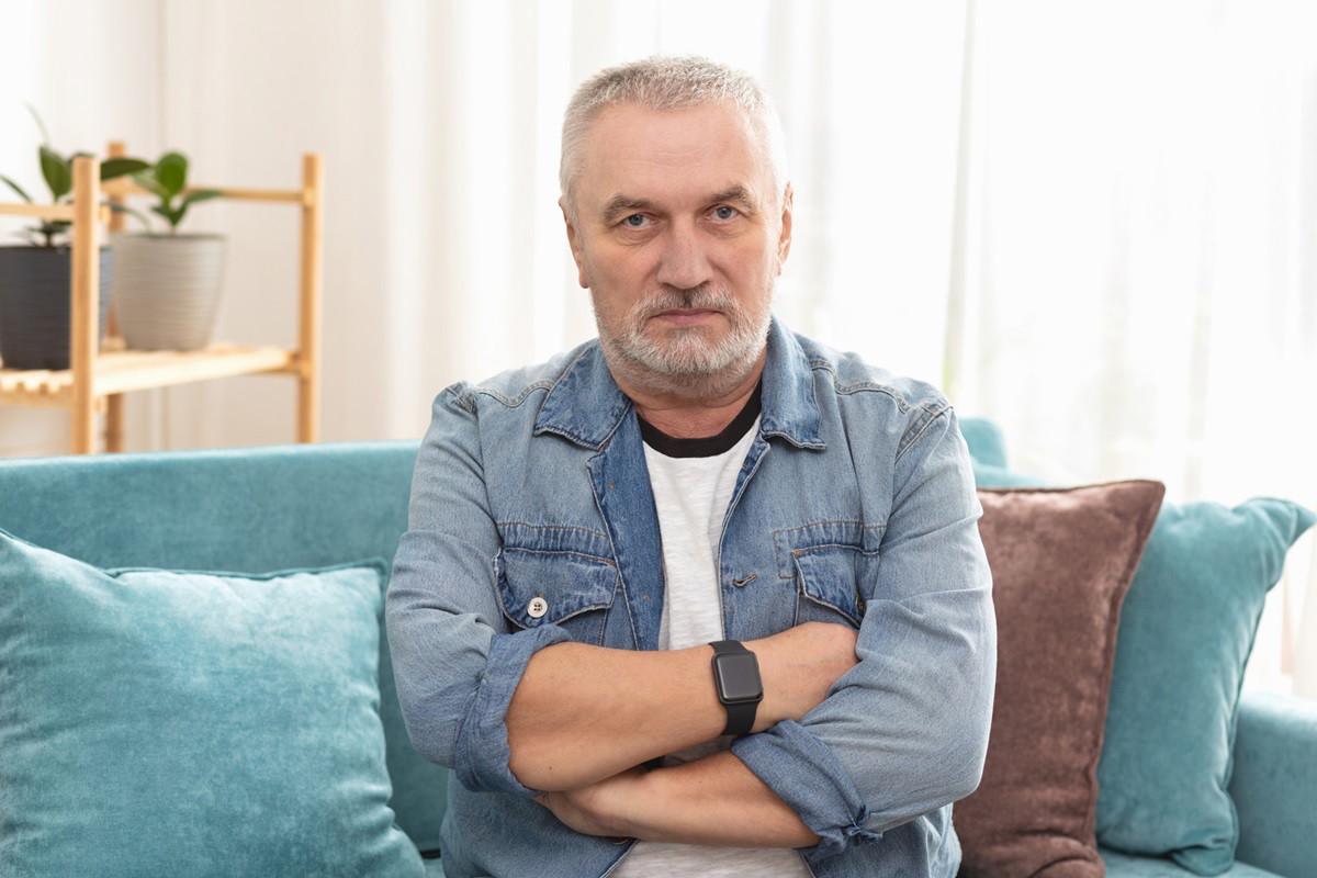 mężczyzna z siwymi włosami i zarostem w wieku około 60 lat w jeansowej koszuli siedzi na kanapie z założonymi rękami i poważną miną