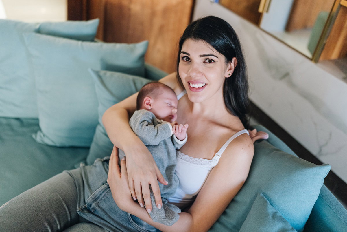 młoda mama w wieku około 30 lat z ciemnymi włosami i w makijażu siedzi na kanapie z noworodkiem i lekko się uśmiecha ale oczy ma smutne