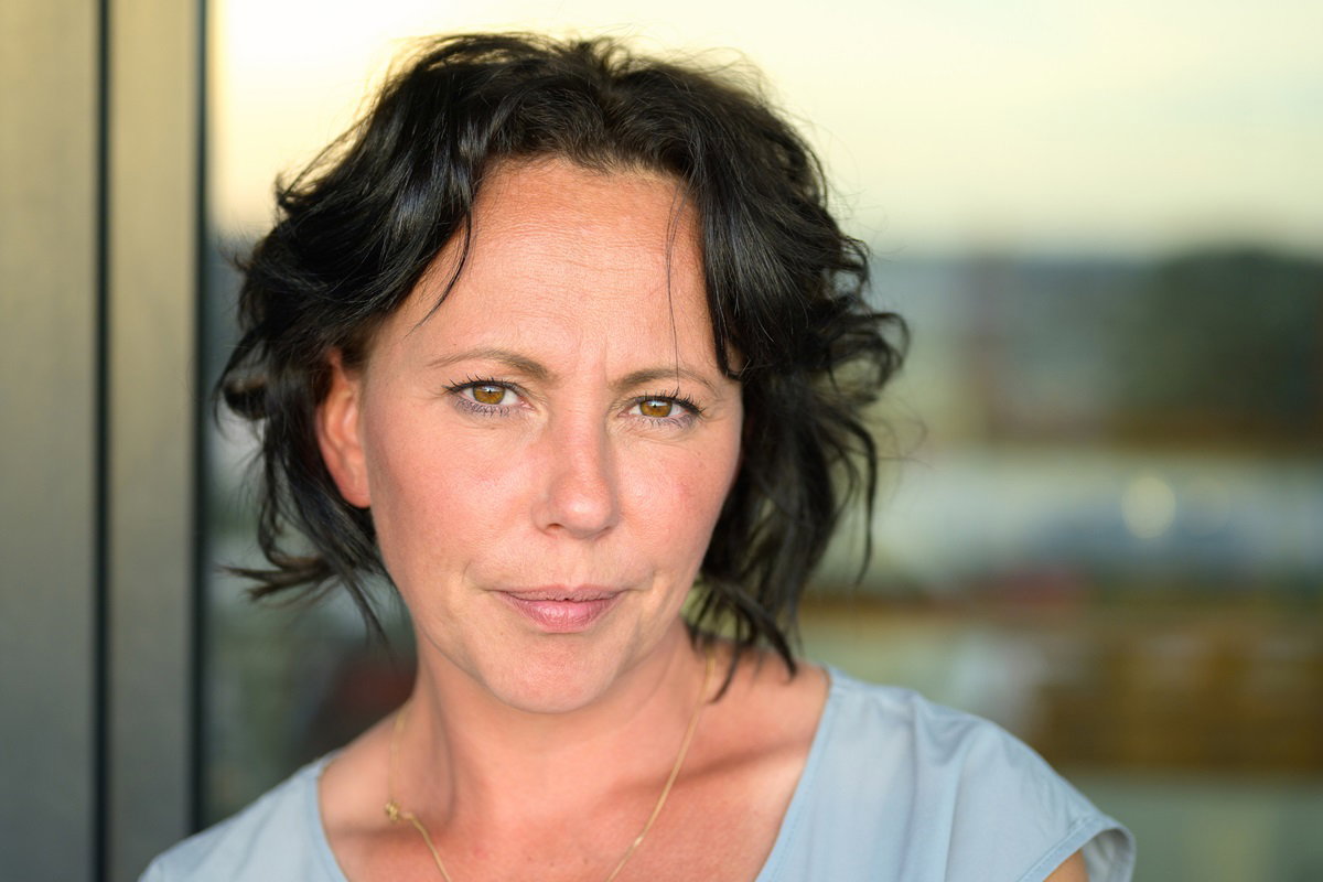 kobieta w średnim wieku około 40 lat z ciemnymi włosami stoi na tle okna w mieście i ma poważną minę