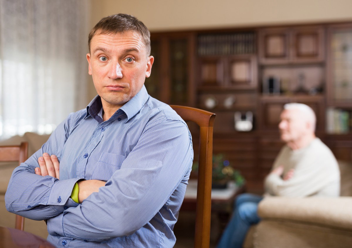 dorosły mężczyzna w wieku około 35 lat w niebieskiej koszuli stoi ze zdziwioną miną na tle meblościanki a w tle siedzi starszy senior