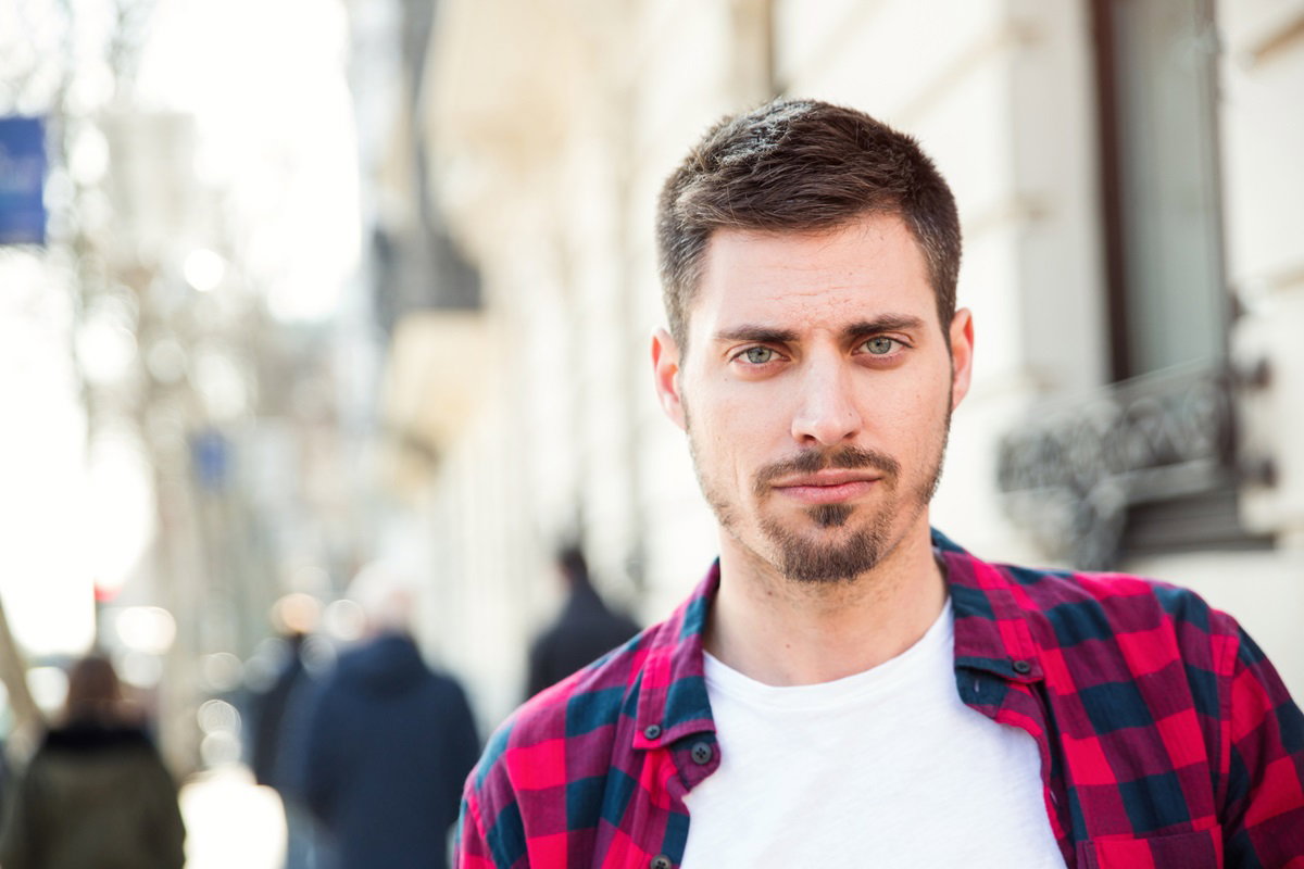 młody mężczyzna w wieku około 30 lat z zarostem i w koszuli czerwonej w kratę stoi na ulicy ze zmartwioną miną
