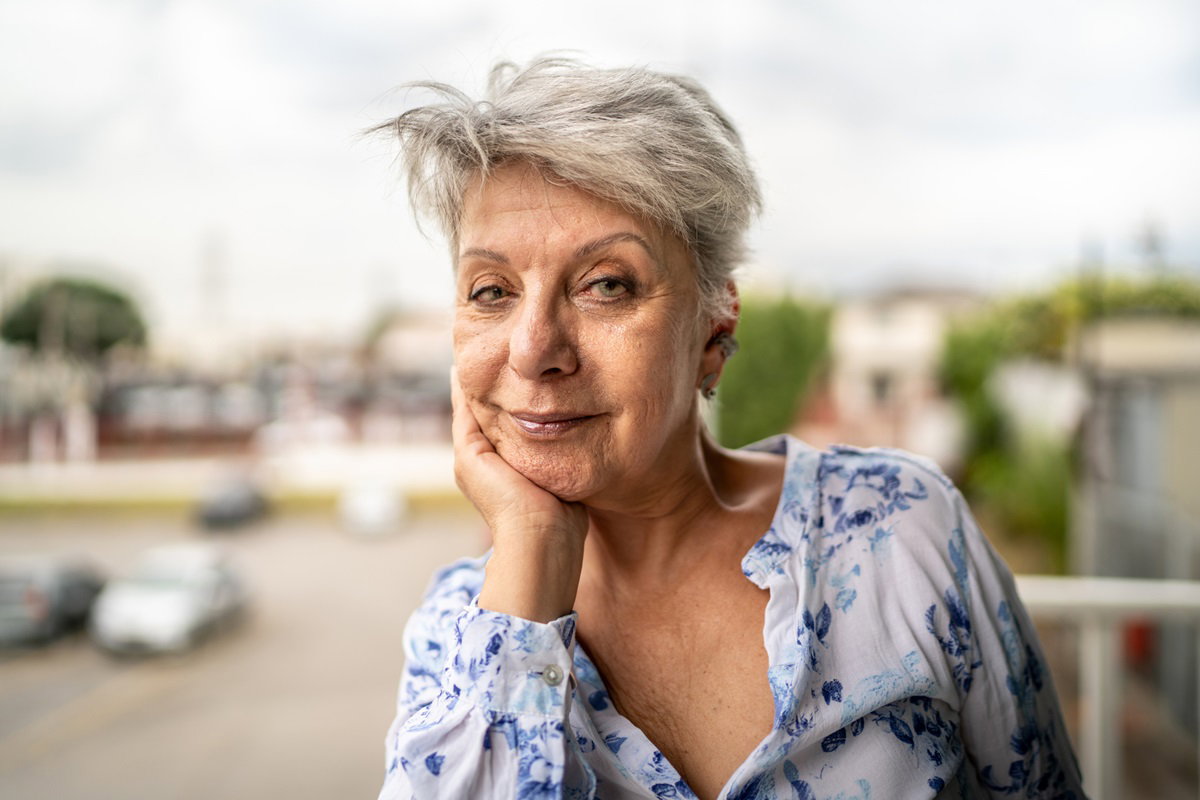 kobieta w wieku około 70 lat z krótkimi siwymi włosami ostrzyżonymi nowocześnie i w bluzce w kwiaty podpiera głowę ręką i patrzy z przekąsem
