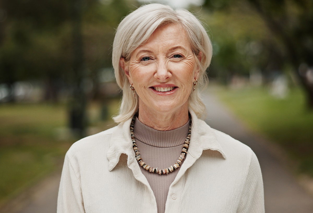 kobieta w wieku senioralnym 60+ z blond włosami i koralami na szyi stoi na drodze na zewnątrz i uśmiecha się promiennie