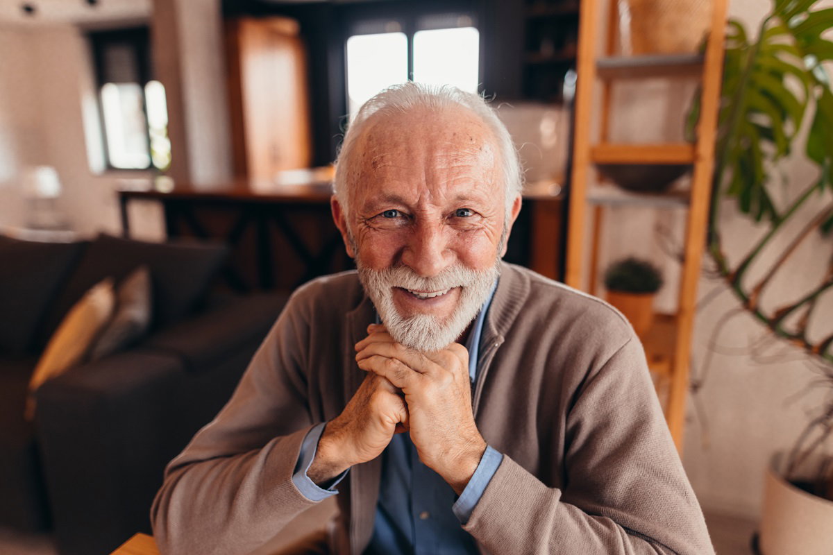 mężczyzna w wieku senioralnym siwy z brodą i wąsami w sweterku siedzi w salonie i uśmiecha się od ucha do ucha