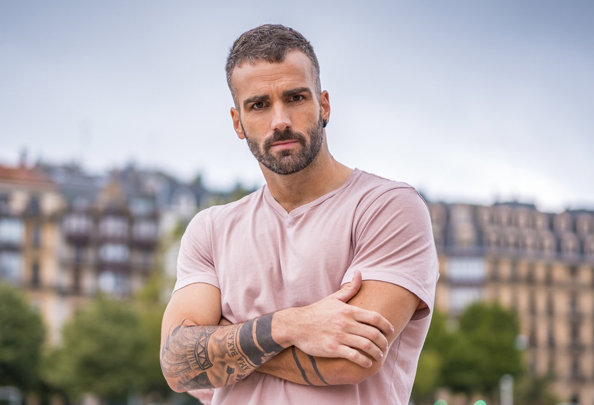 przystojny mężczyzna wieku około 30 lat z zarostem i tatuażami w różowym t-shircie stoi z założonymi rękami i poważną miną