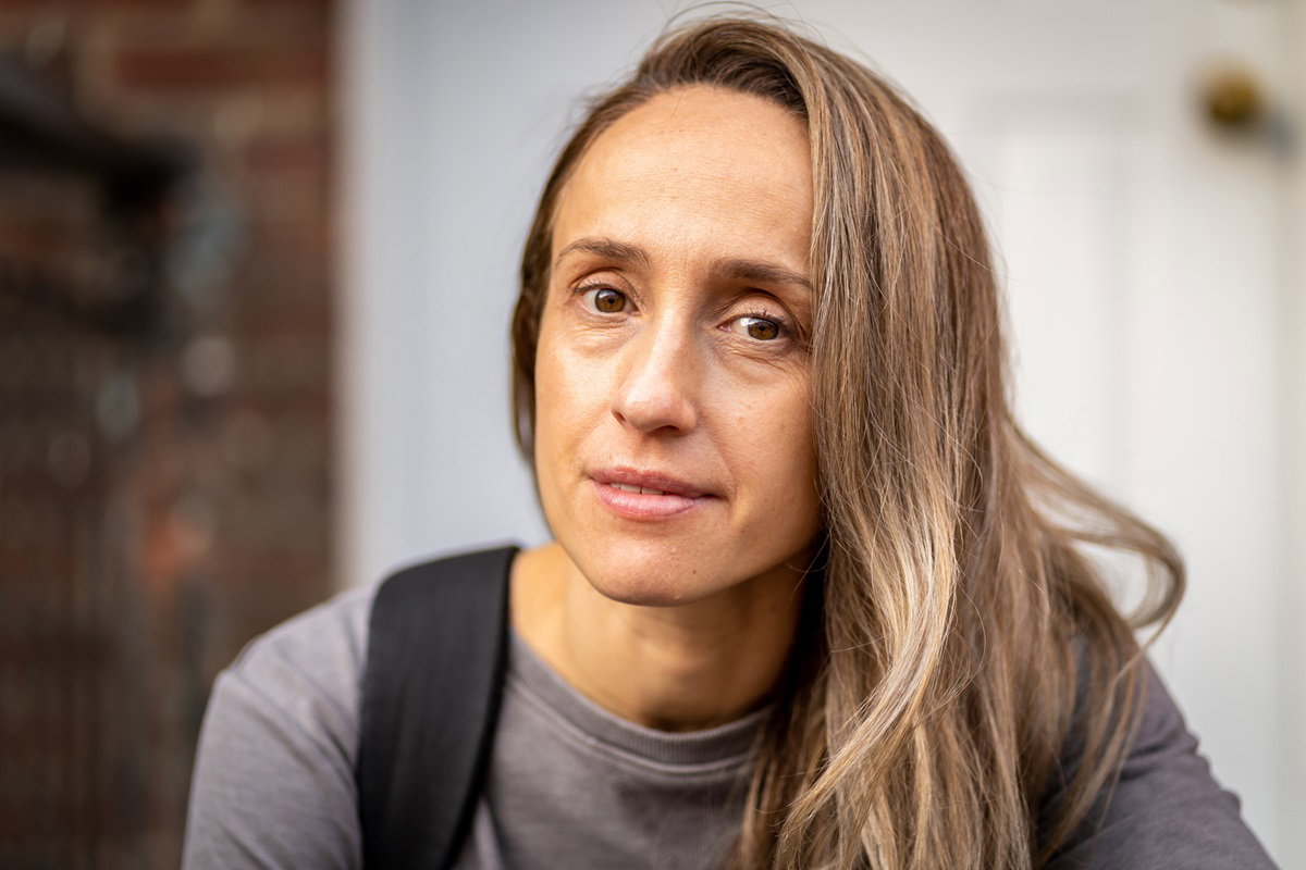 kobieta w wieku około 40 lat z długimi włosami i z plecakiem na plecach siedzi na zewnątrz na schodach i lekko się uśmiecha