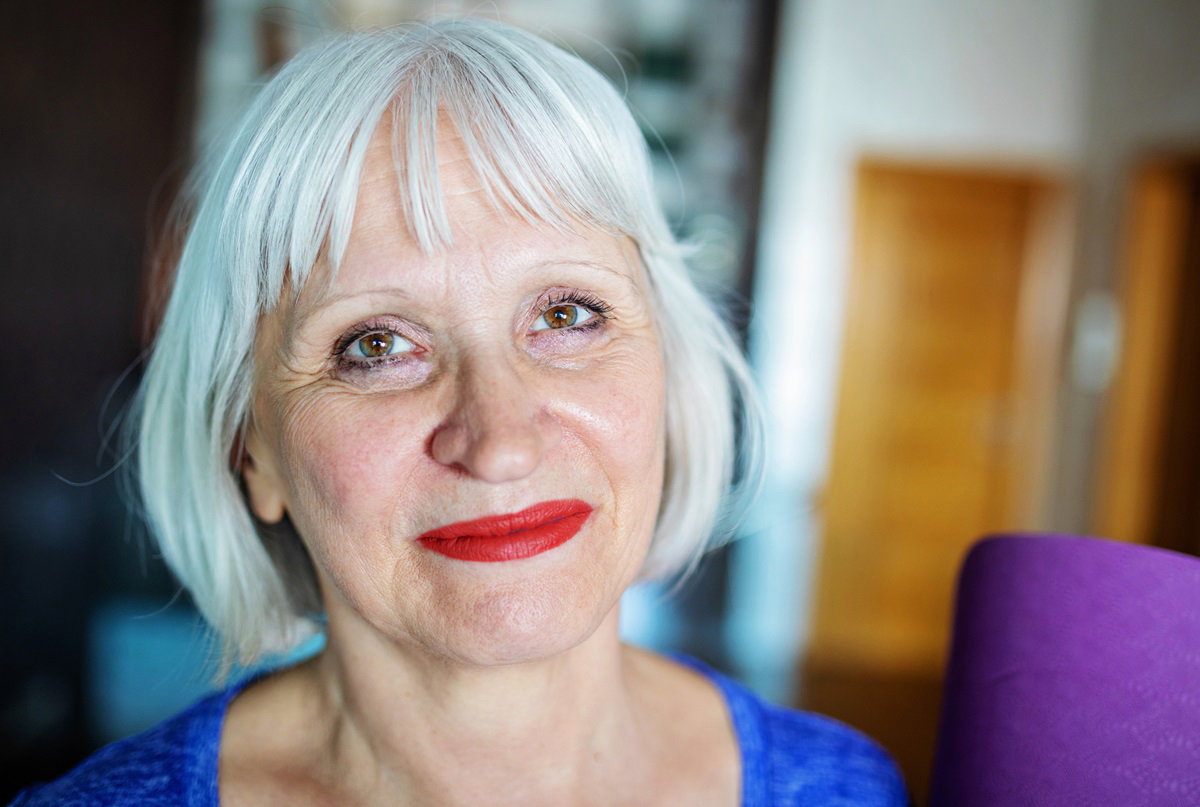 kobieta w wieku około 65 lat z bardzo jasnymi włosami na pazia z grzywką i ustami pomalowanymi na czerwono siedzi w mieszkaniu i uśmiecha się