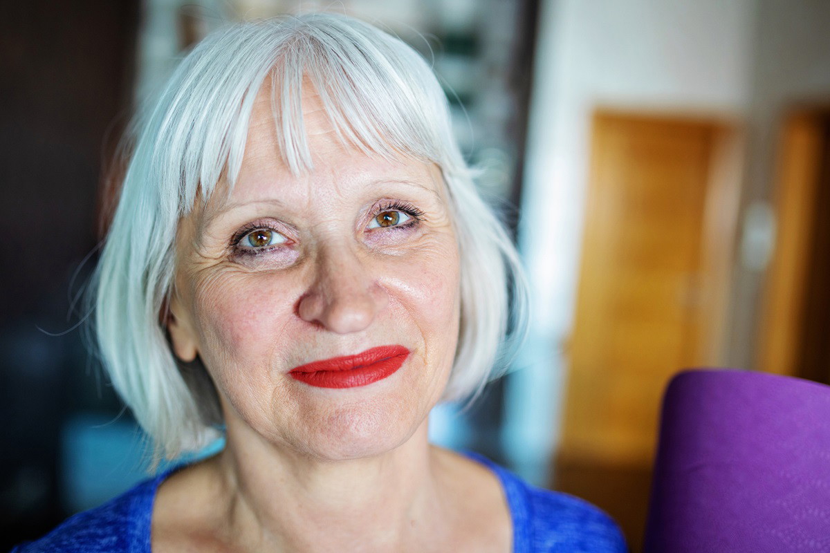 kobieta w wieku około 65 lat z bardzo jasnymi włosami na pazia z grzywką i ustami pomalowanymi na czerwono siedzi w mieszkaniu i uśmiecha się