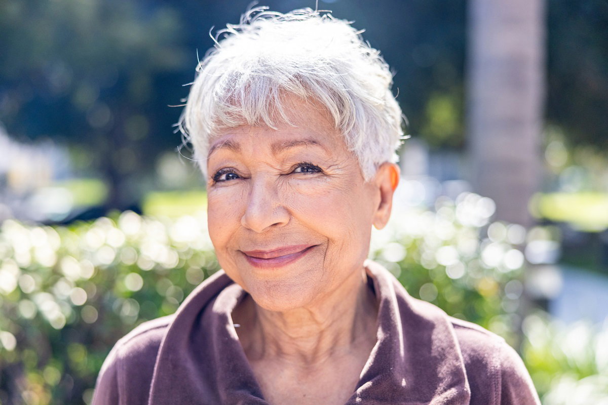 kobieta w senioralnym wieku 70+ z krótkimi siwymi włosami siedzi na zewnątrz w słońcu i uśmiecha się zadziornie