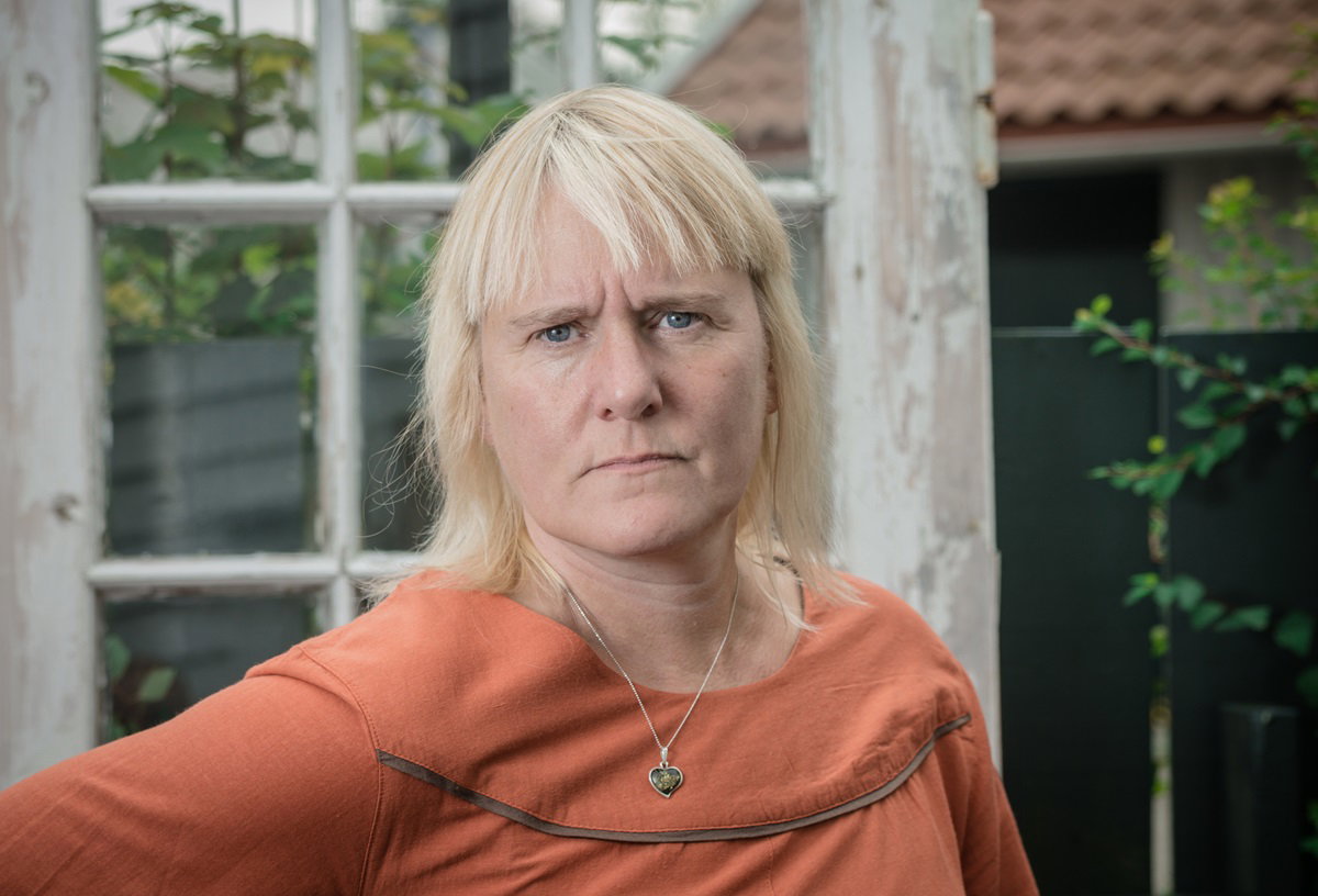 kobieta w wieku około 65 lat w pomarańczowej bluzce i z blond fryzurą z grzywką stoi na tle ramy i domu z zagniewaną miną