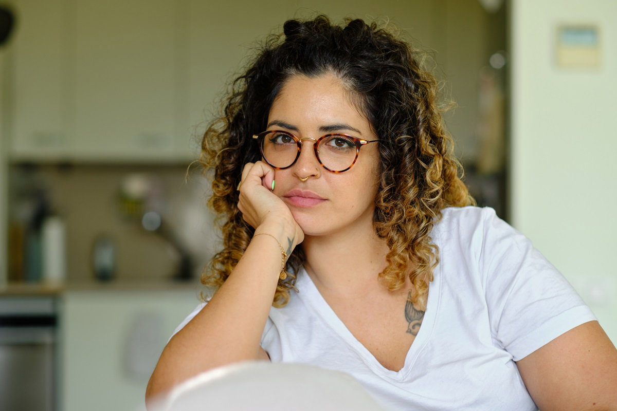 młoda kobieta z kręconymi włosami i w okularach z kolczykiem w nosie siedzi zdegustowana i smutna w kuchni