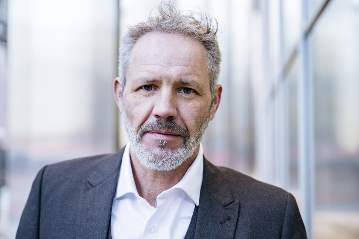 przystojny mężczyzna w wieku około 50 lat siwy z zarostem w marynarce i koszuli stoi na tle budynku i patrzy poważnie