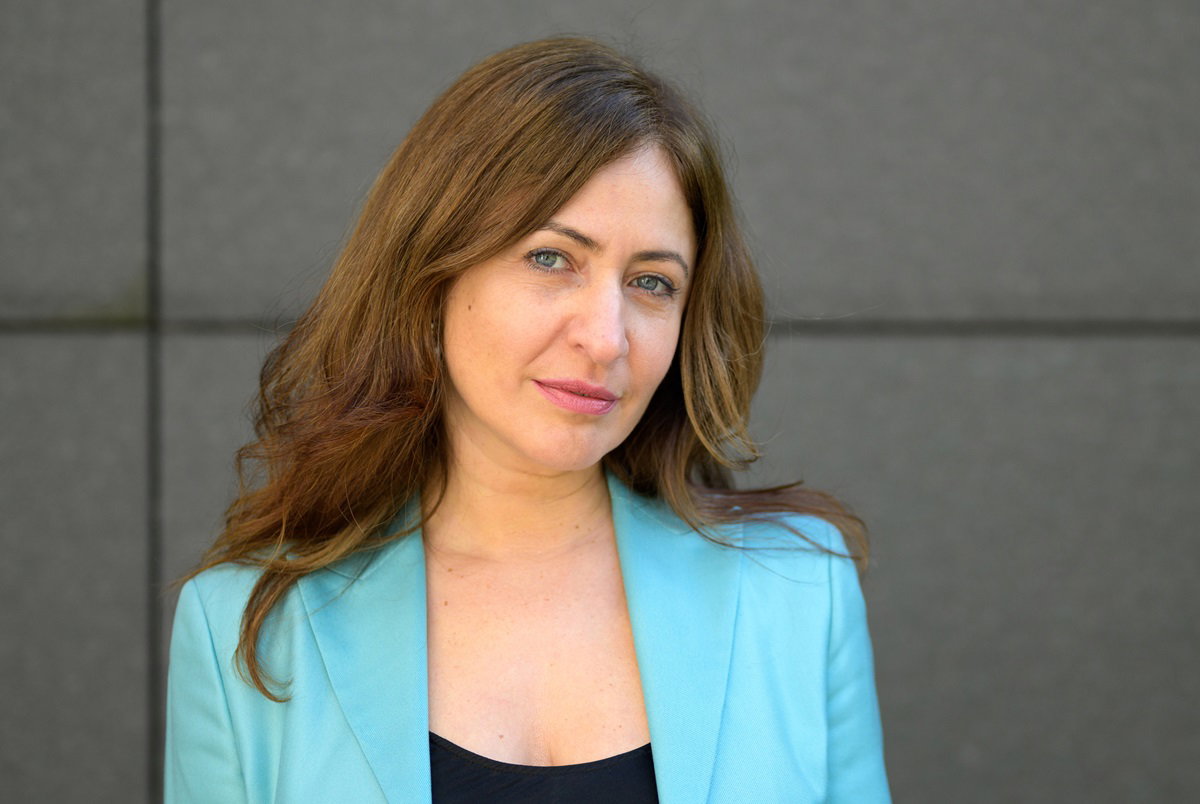 kobieta w wieku około 35 lat w błękitnej marynarce z długimi włosami stoi na tle ściany budynku i patrzy przed siebie