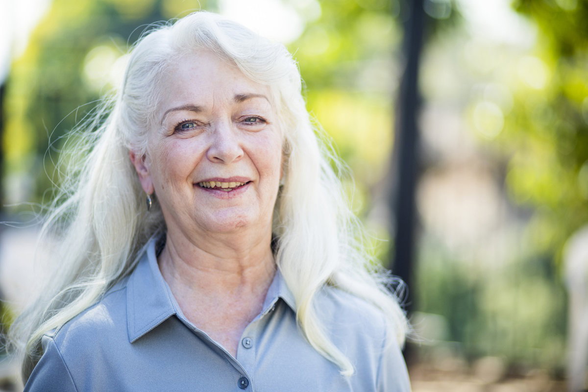 kobieta w wieku 60+ z siwymi długimi włosami i w niebieskiej koszuli stoi na zewnątrz i uśmiecha się z przekąsem