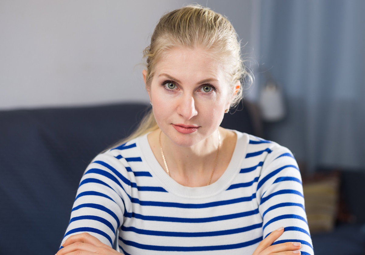 kobieta w wieku około 40 lat ze spiętymi blond włosami i w bluzce w paski siedzi zmartwiona z założonymi rękami