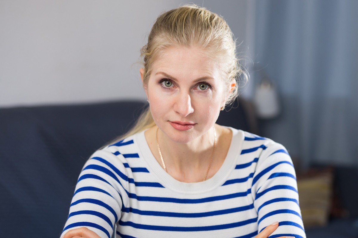 kobieta w wieku około 40 lat ze spiętymi blond włosami i w bluzce w paski siedzi zmartwiona z założonymi rękami