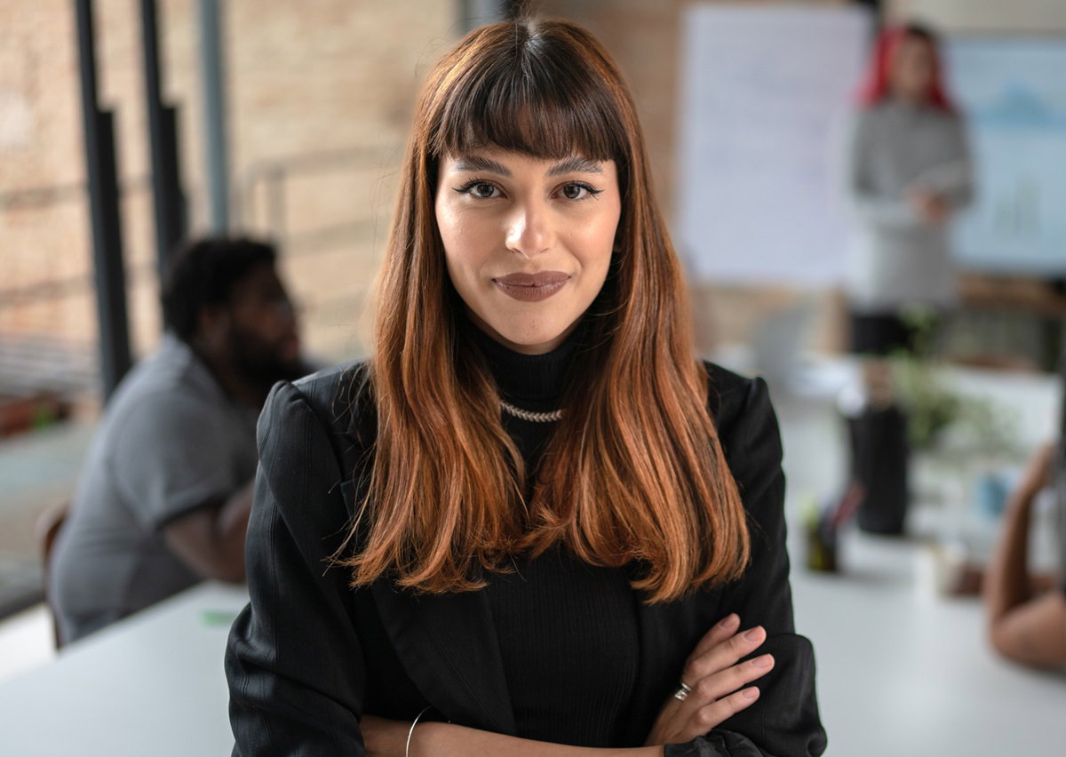 młoda dziewczyna z rudymi długimi włosami z grzywką stoi w agencji marketingowej z rękami złożonymi i uśmiechem