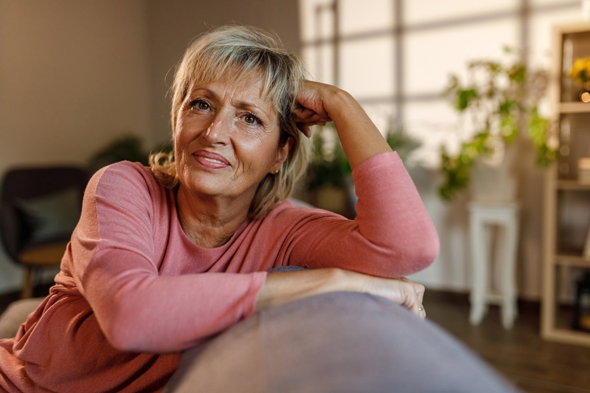 kobieta w dojrzałym wieku 50 plus w różowej bluzce z krótkimi blond włosami siedzi na kanapie i rozmyśla