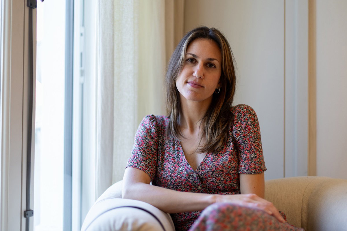 młoda kobieta w wieku około 30 lat z długimi włosami siedzi na fotelu w sukience w drobny wzór z lekko kpiącą miną