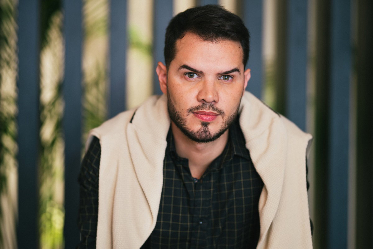 młody mężczyzna w wieku 35 lat z ciemnymi włosami i zarostem z zarzuconym swetrem na ramiona stoi w biurze i patrzy zagniewanym wzrokiem