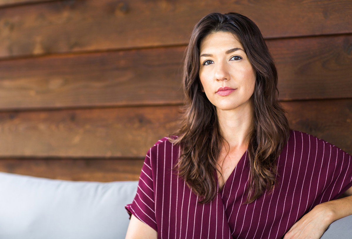 młoda kobieta w wieku około 35 lat z długimi ciemnymi włosami siedzi na tle drewnianej ściany i patrzy poważnym wzrokiem