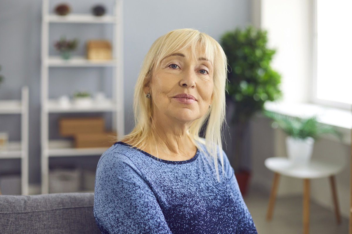 kobieta blondynka w wieku około 60 lat w niebieskiej bluzce i w makijażu siedzi w salonie z zamyśloną miną