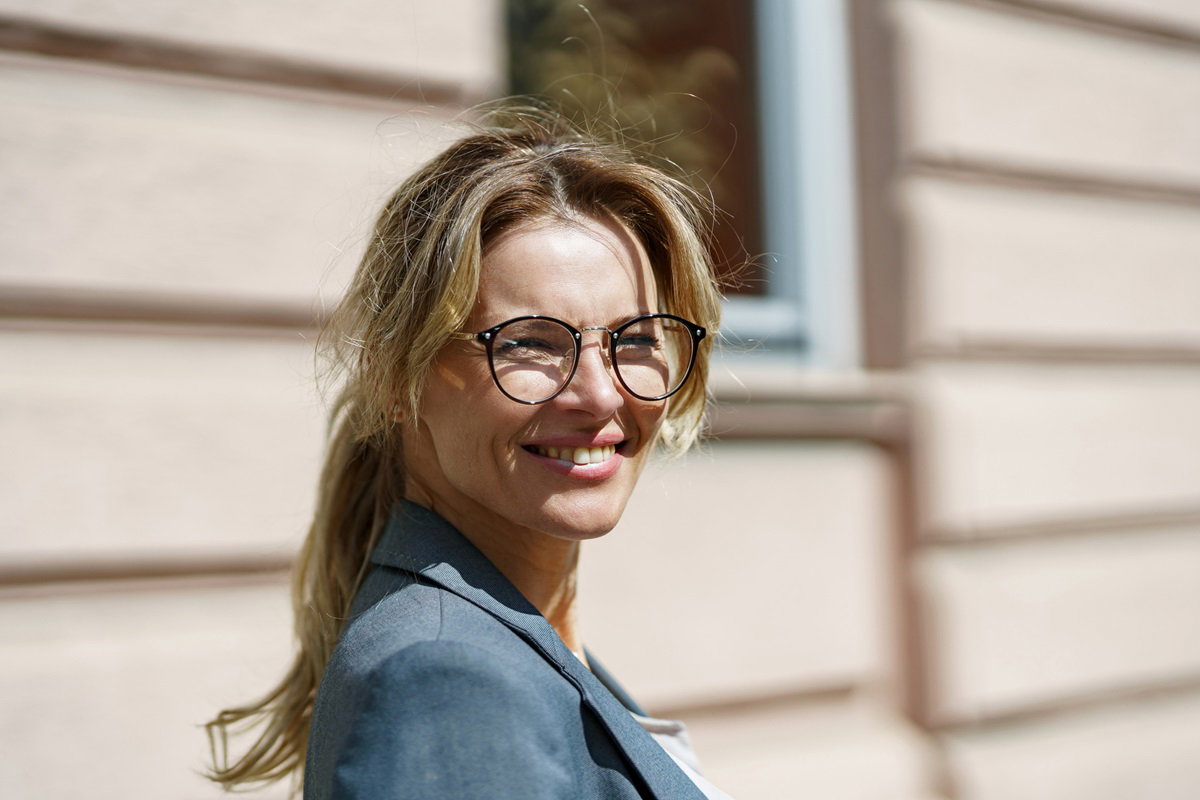 młoda kobieta blondynka w marynarce i w okularach stoi na tle budynku w słońcu i się uśmiecha