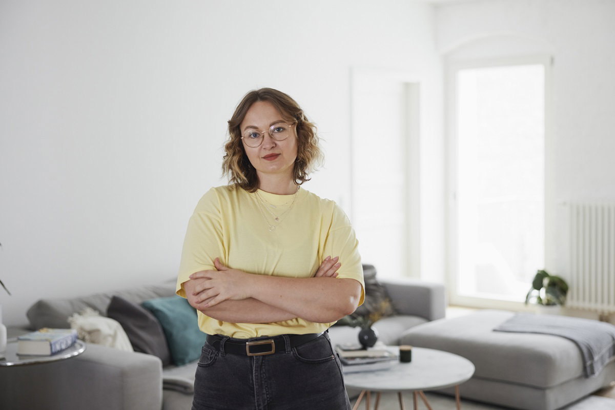 kobieta w żółtym t-shircie i w okularach stoi w salonie na tle kanapy z założonymi rękami