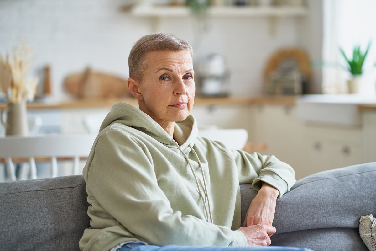 kobieta w wieku 55+ w bluzie z kapturem z krótkimi włosami siedzi na kanapie z podejrzliwą miną