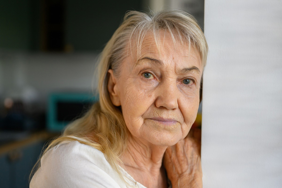kobieta seniorka w wieku 65+ z siwiejącymi blond długimi włosami stoi oparta o framugę ze smutną miną i bólem w oczach
