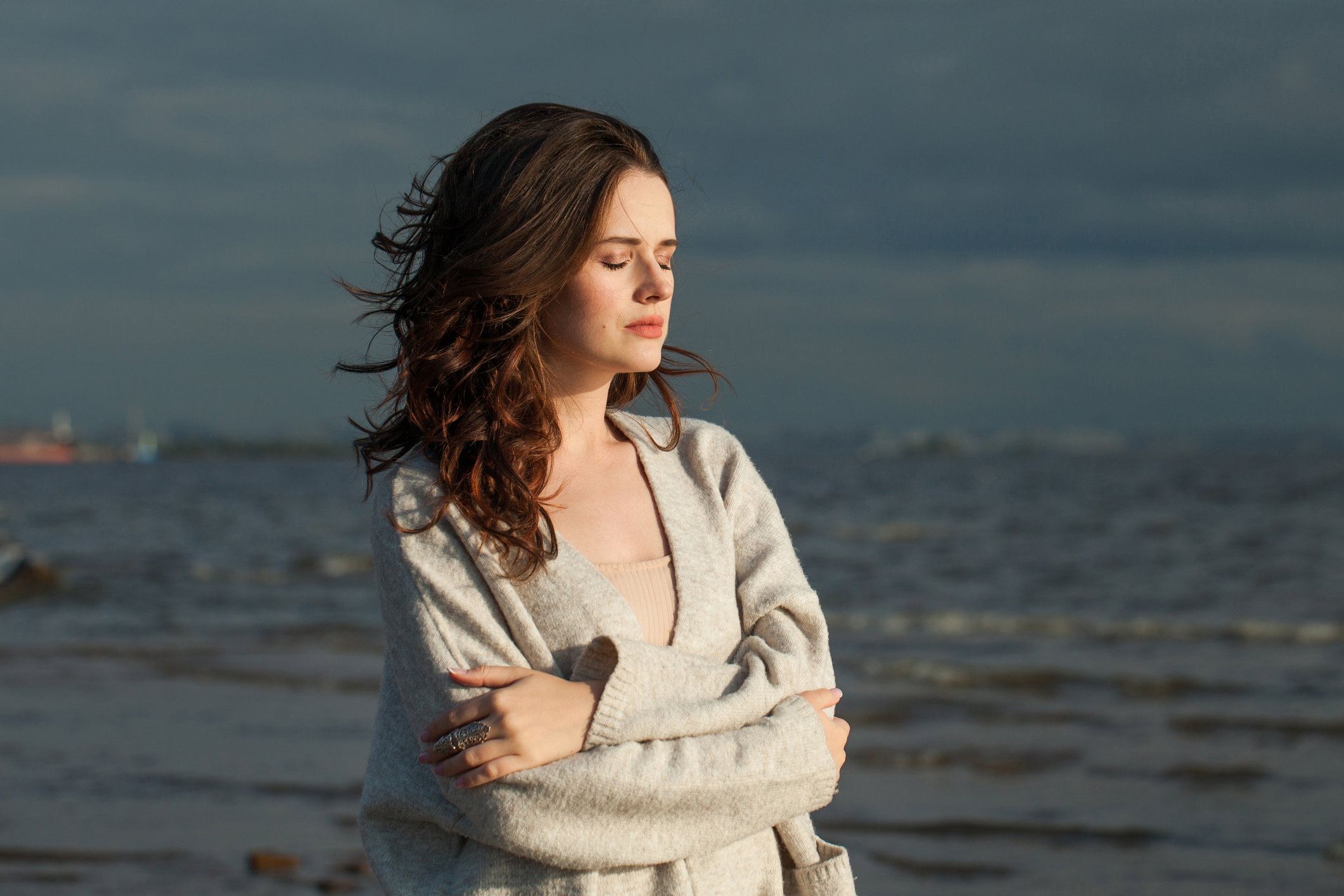 Resting young lady brunette model on sea beach. Beauty woman with makeup and long dark wavy hairstyle outdoors. Tranquility emotion mood, travel, season and holiday concept