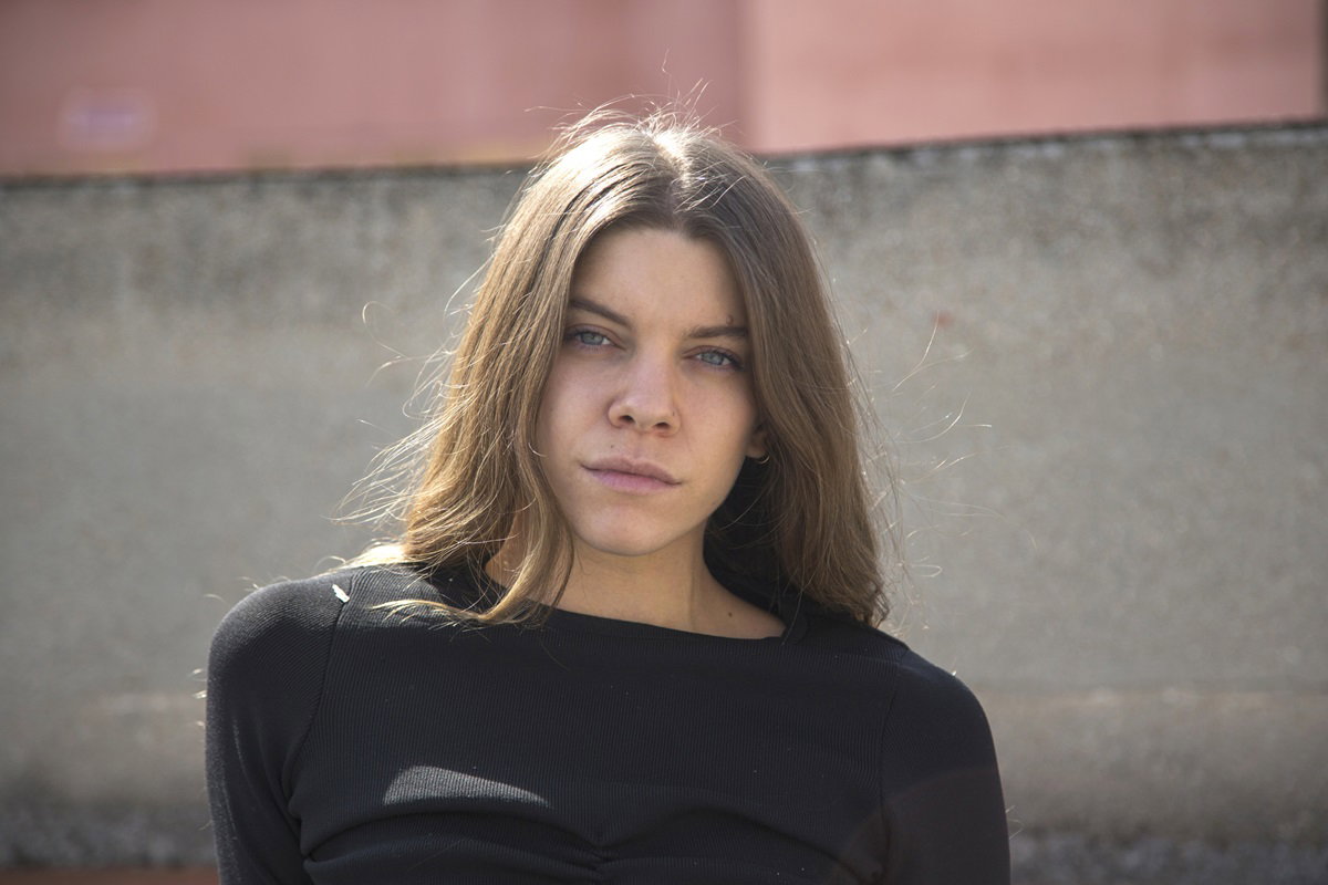 młoda dziewczyna w wieku około 30 lat w czarnym crop topie z długimi włosami siedzi na tle muru w słońcu i ma zamyśloną minę