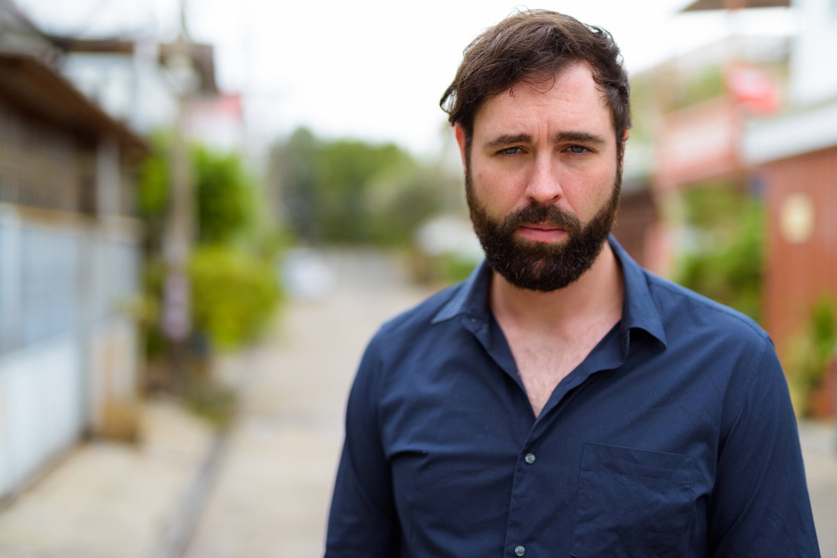 młody mężczyzna  w wieku około 35 lat z brodą i wąsami w granatowej koszuli stoi na zewnątrz ze smutną miną