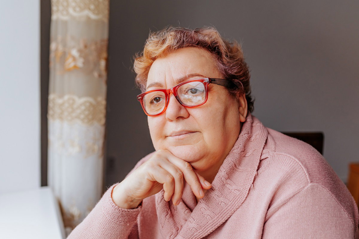 kobieta w dojrzałym wieku około 65+ w okularach i różowym swetrze siedzi i patrzy w okno zamyślona