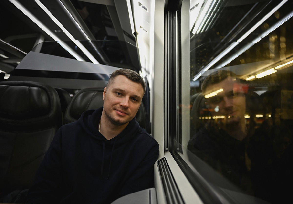 młody mężczyzna siedzi w pociągu przy oknie i lekko się uśmiecha zamyślony