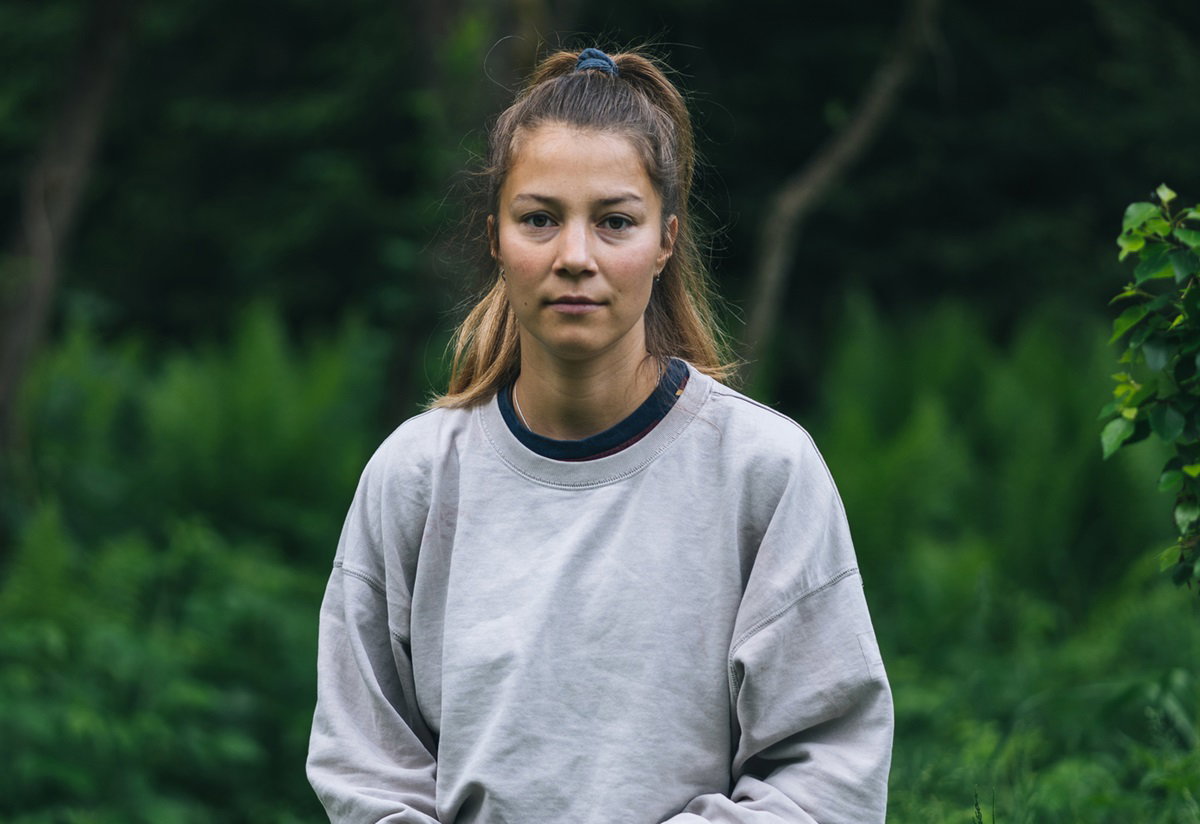 młoda kobieta w szarej bluzie na tle zieleni siedzi zasmucona