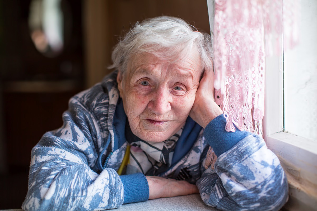 seniorka 80+ z siwymi włosami siedzi przy stole koło okna w bluzie ze zmartwioną miną