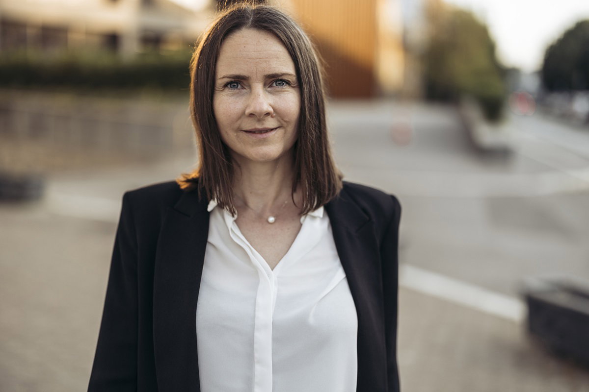 kobieta w dojrzałym wieku w marynarce i koszuli stoi ze smutnym uśmiechem na ulicy