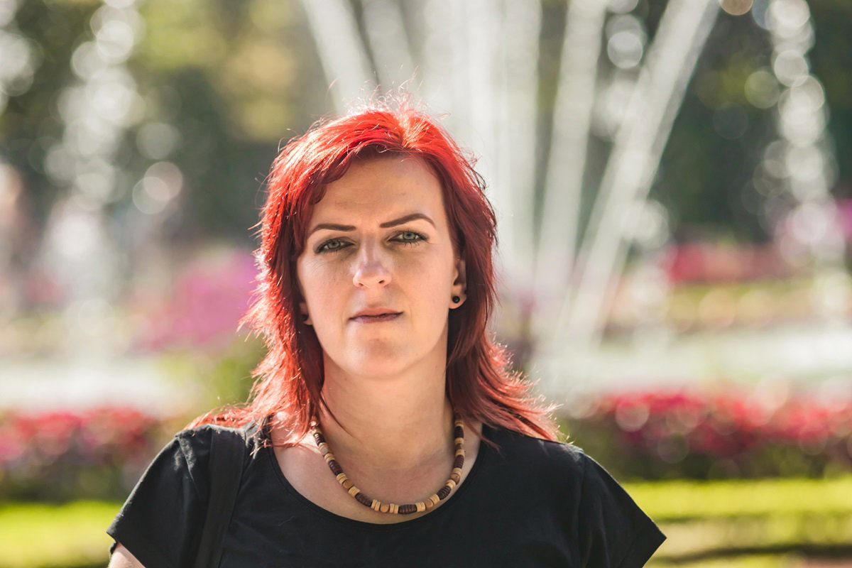 młoda kobieta z pofarbowanymi włosami na intensywny kolor siedzi na tle fontanny na rynku ze skwaszoną miną